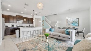 A modern kitchen with dark wooden cabinets, a central island, and a dining area with a white sofa and a glass coffee table. - Spacious 5BR Villa w/ Private Pool – near Disney! (Kissimmee)