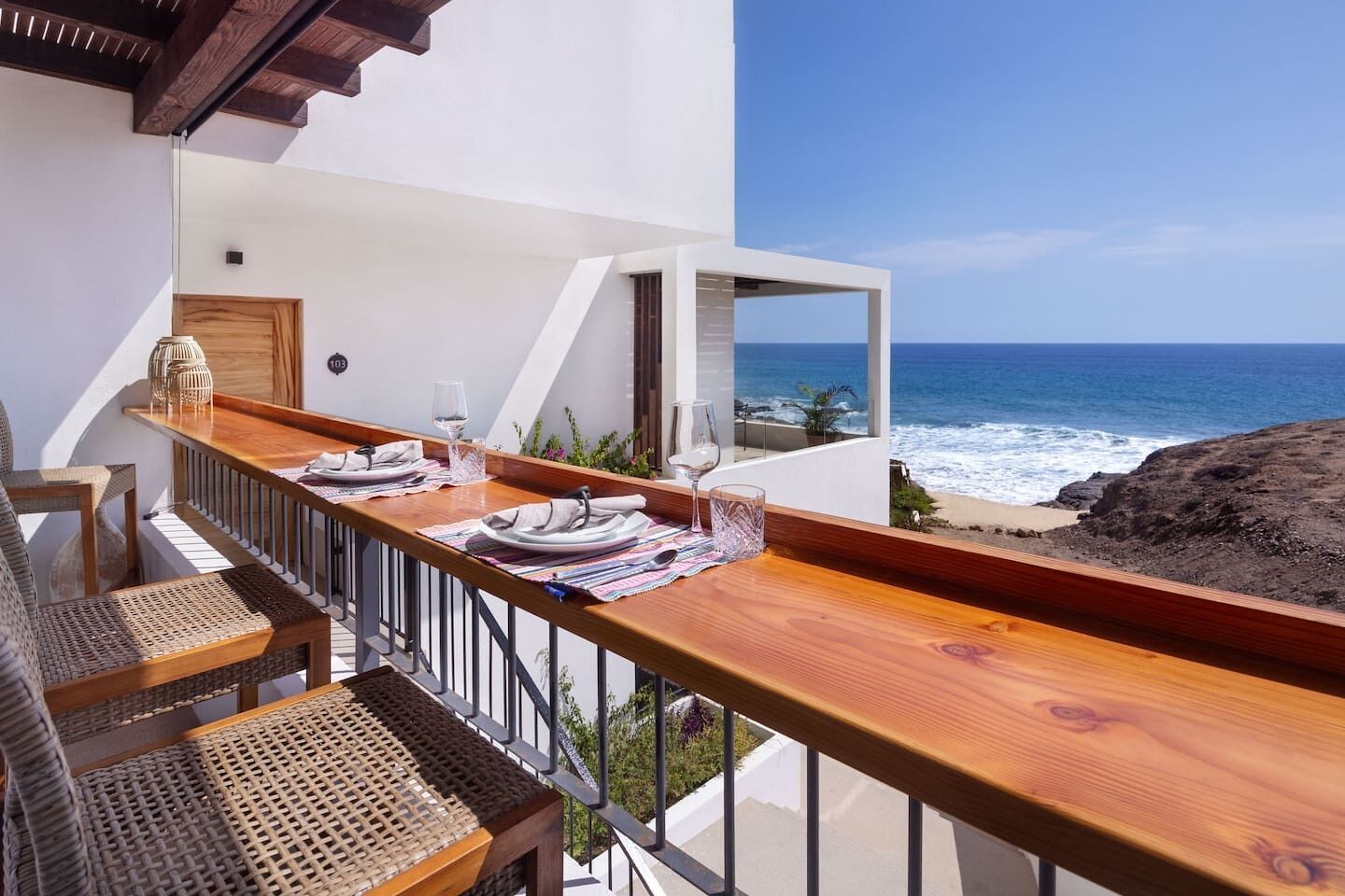 A balcony with a wooden table set for dining, overlooking a beach with the ocean and rocky terrain.