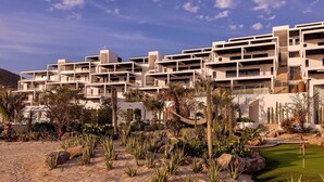 Hotel moderno com várias varandas, um jardim ajardinado com cactos e palmeiras, e área de piscina.