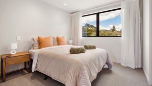 A bedroom with a bed, nightstand, and window with curtains. - Unwind and Relax in Central Waihi Beach (#B4) (Waihi Beach)