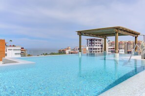 A clear swimming pool with a wooden pergola, surrounded by multi-story buildings with balconies, under a clear sky. - Romantic Zone Luxury | V399 Condo w/ Infinity Pool (Puerto Vallarta)