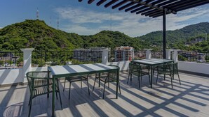 A rooftop terrace with a pergola, tables, and chairs, overlooking a green hillside with buildings in the distance. - Condo Luz Romantic Zone with Rooftop Infinity Pool, Hot Tub (Puerto Vallarta)