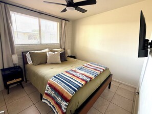 Un dormitorio con una cama, una mesita de noche, un ventilador de techo, una ventana con persianas y un televisor montado en la pared.