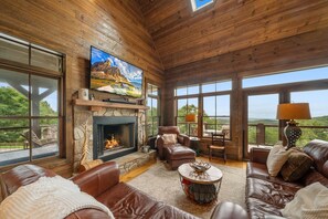 Ein großzügiges Wohnzimmer mit Kamin, Ledersofas, einem Fernseher und großen Fenstern mit Blick ins Freie.