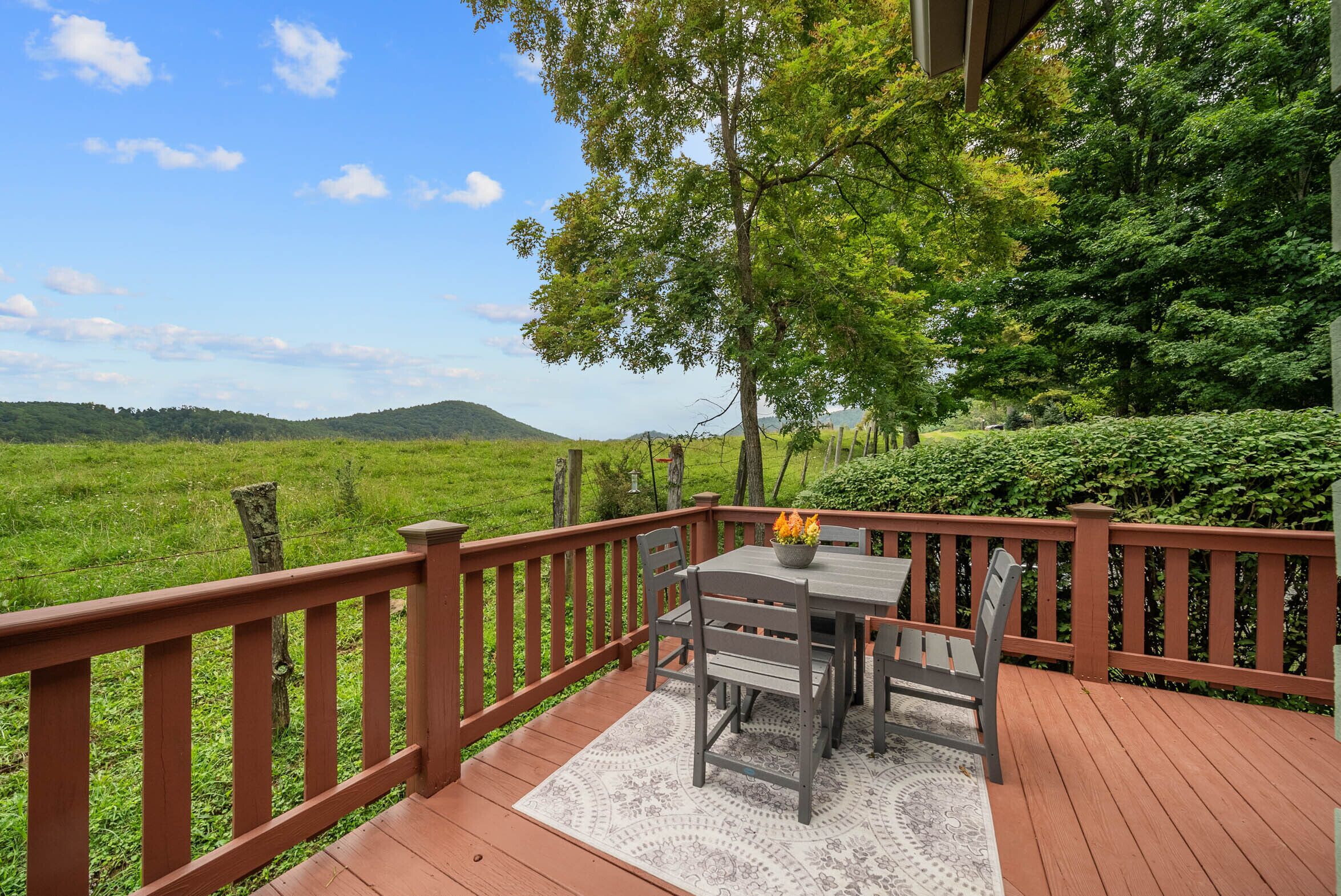 A wooden deck with a table and chairs, surrounded by lush greenery and a view of rolling hills.