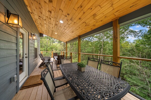 A covered outdoor patio with a wooden ceiling, a metal table with chairs, and a view of the surrounding forest.