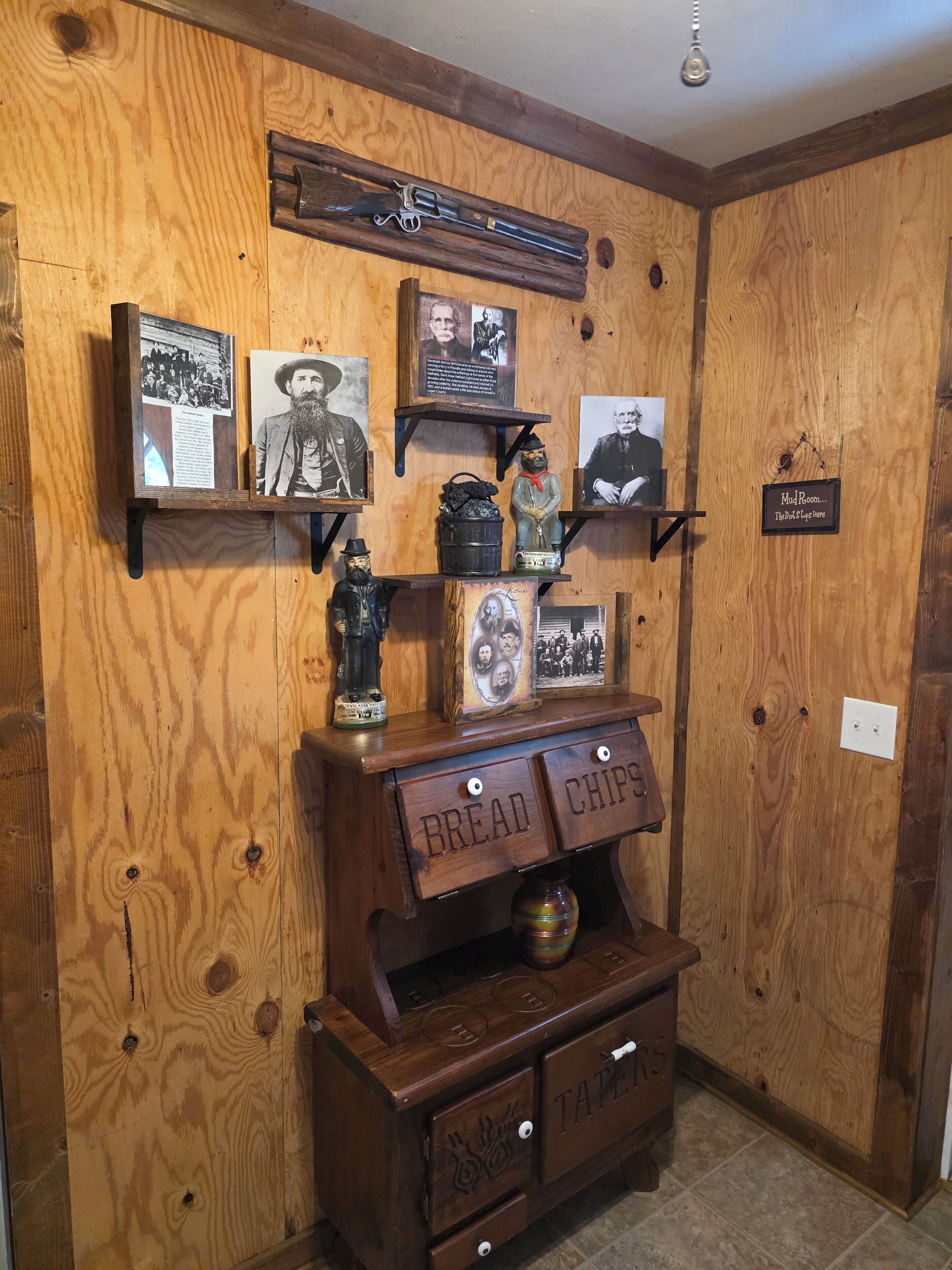 A wooden wall with shelves displaying historical photos, a revolver, and a sign reading "Mud Room... The Dude's Log Cabin."