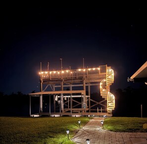 Une structure en bois à plusieurs niveaux, dotée d’un escalier en colimaçon, d’une enceinte en verre et d’un chemin pavé éclairé.
