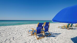 Uma praia com areia branca e águas azul-claras. Há cadeiras de praia e um guarda-sol azuis.