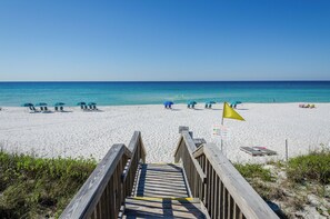 A wooden boardwalk leading to a beach with white sand and clear blue water. There are several beach umbrellas and lounge chairs along the shore.