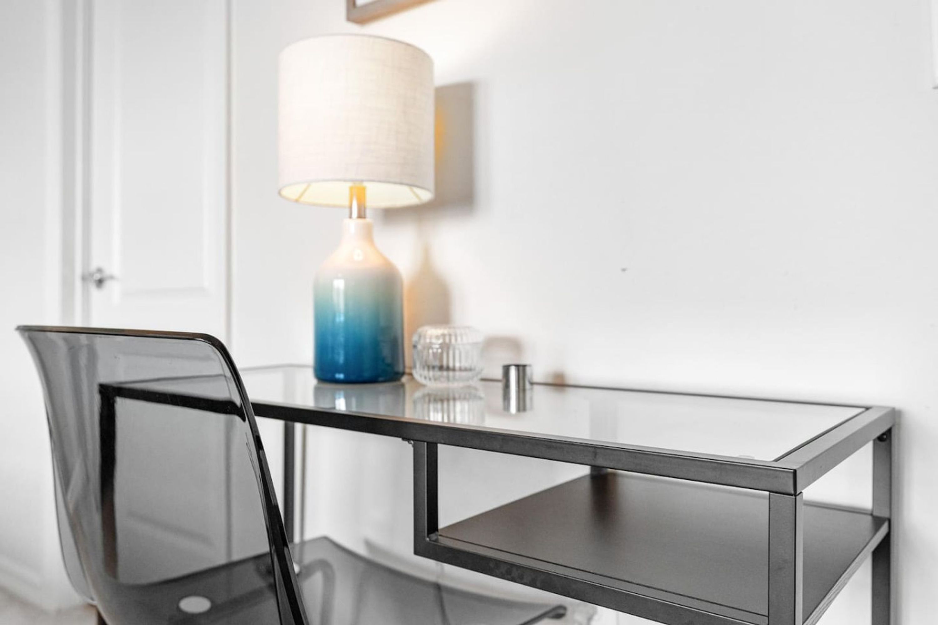 Une table moderne en verre avec un cadre en métal, une chaise transparente, une lampe bleue et blanche, et un petit objet en verre posé sur la table.