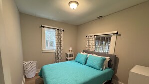 A bedroom with a bed, two windows with patterned curtains, a bedside table with a lamp, and a white trash bin. - Located near UGA, Normaltown, & Downtown Athens. Charming 2-bed cottage w/ AC. (Athens)
