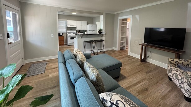 A modern living room with a grey sofa, a flat-screen TV, and a kitchen in the background. - Located near UGA, Normaltown, & Downtown Athens. Charming 2-bed cottage w/ AC. (Athens)