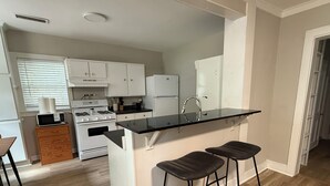 A modern kitchen with white cabinets, a black countertop, and a built-in stove. There are two bar stools and a window with blinds. - Located near UGA, Normaltown, & Downtown Athens. Charming 2-bed cottage w/ AC. (Athens)