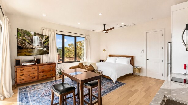 Une chambre à coucher comprenant un lit, un bureau avec deux chaises, une télévision, un ventilateur de plafond et une grande fenêtre avec des rideaux.