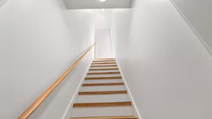 Un escalier avec des marches en bois et une rampe métallique, qui monte dans un couloir étroit aux murs blancs.