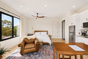 A modern hotel room with a bed, a dining table, a sofa, a ceiling fan, and a kitchen area.