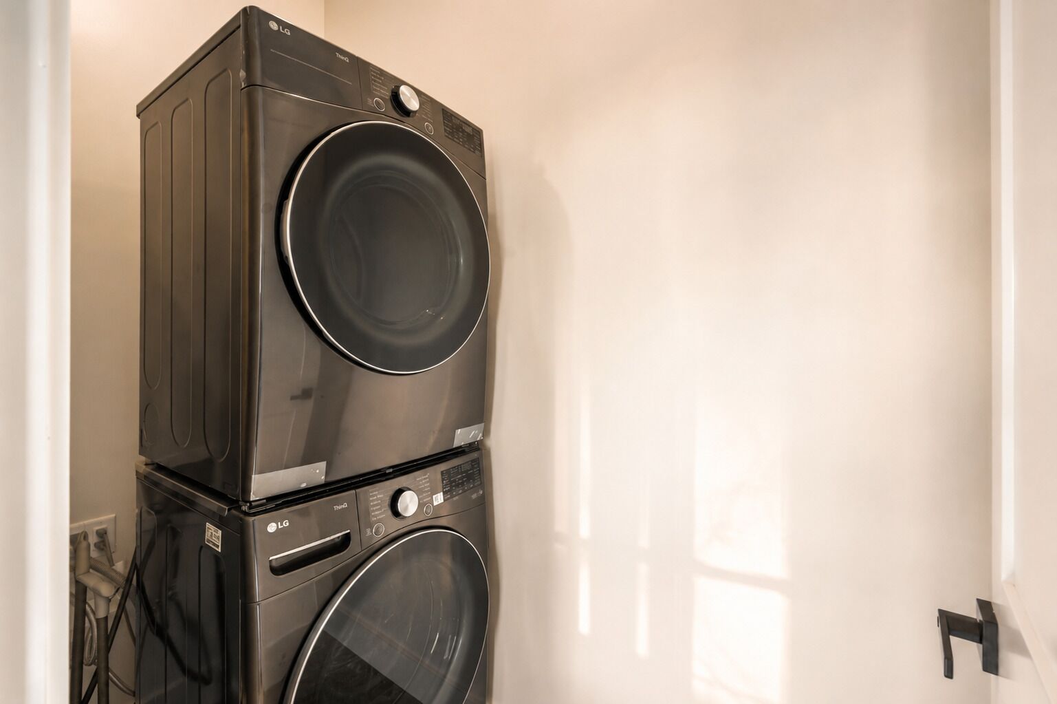 Un appareil combiné lave-linge et sèche-linge, de type « superposé », se trouve dans une pièce aux murs blancs, avec une porte.