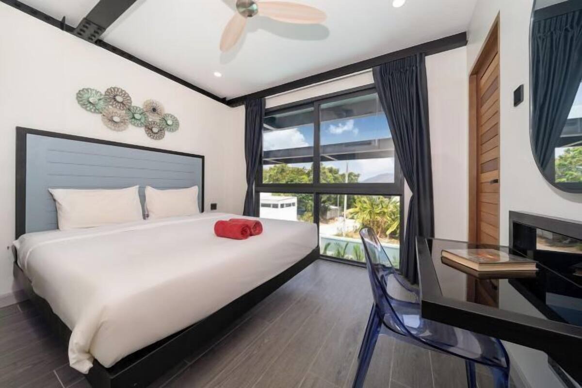 A modern hotel room with a bed, a desk, a chair, a ceiling fan, and a large window with curtains.