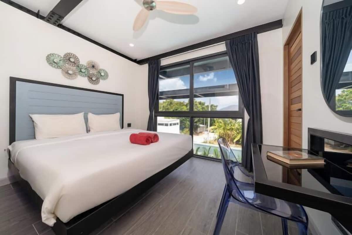A modern hotel room with a bed, a desk, a chair, a ceiling fan, and a large window with curtains.