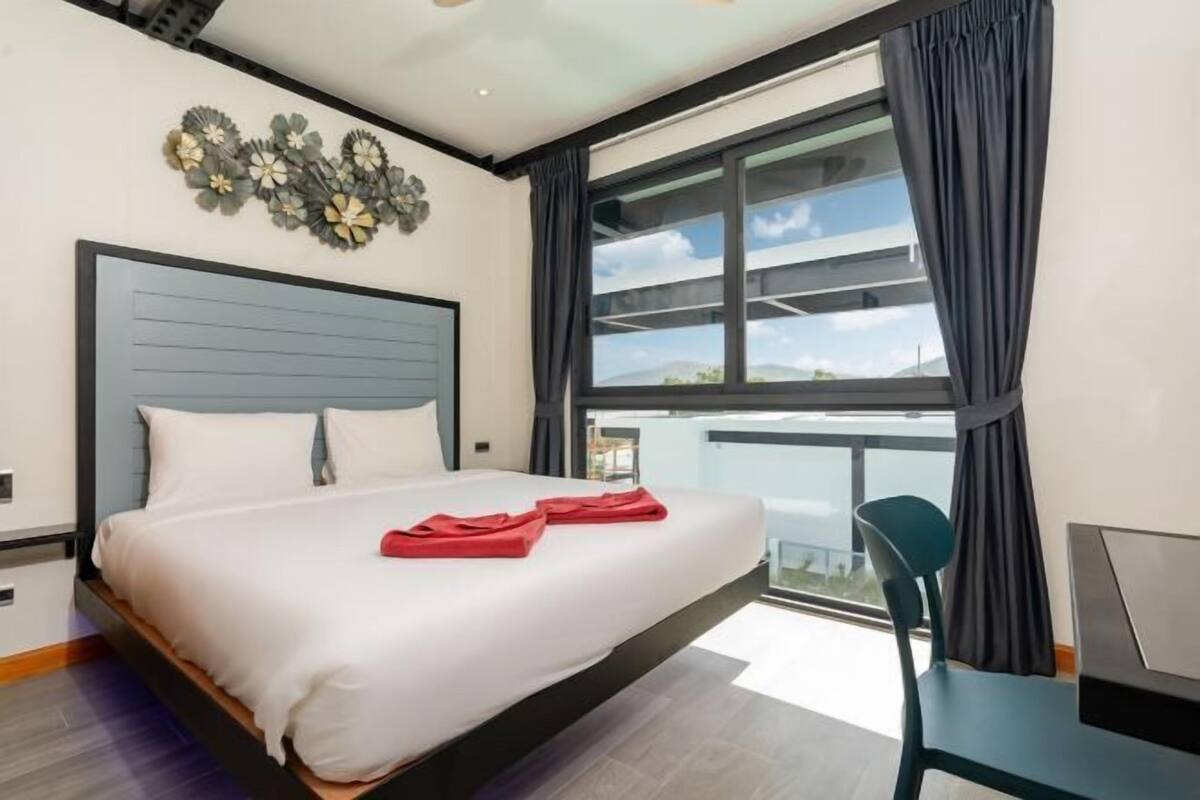 A modern hotel room with a bed, a desk, a chair, a large window with curtains, and a decorative wall piece.