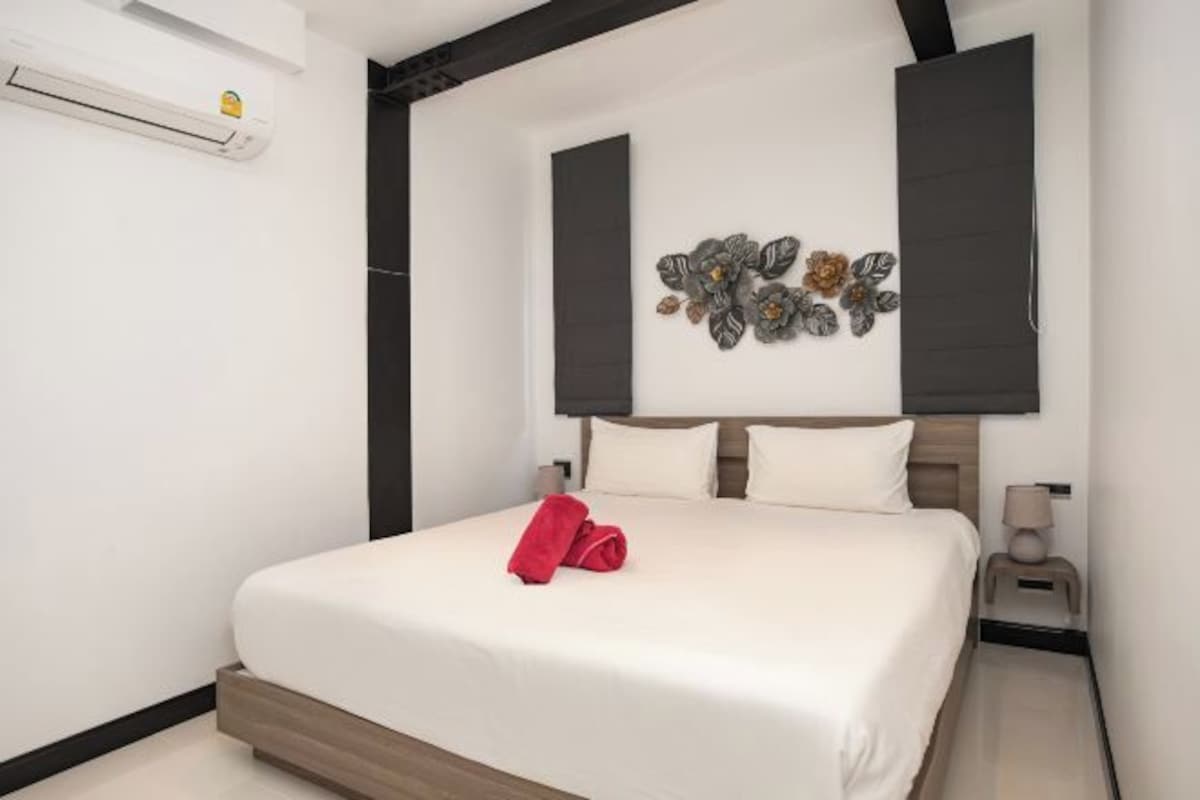 A neatly made bed with a red towel, a wall-mounted air conditioner, and a decorative wall piece.