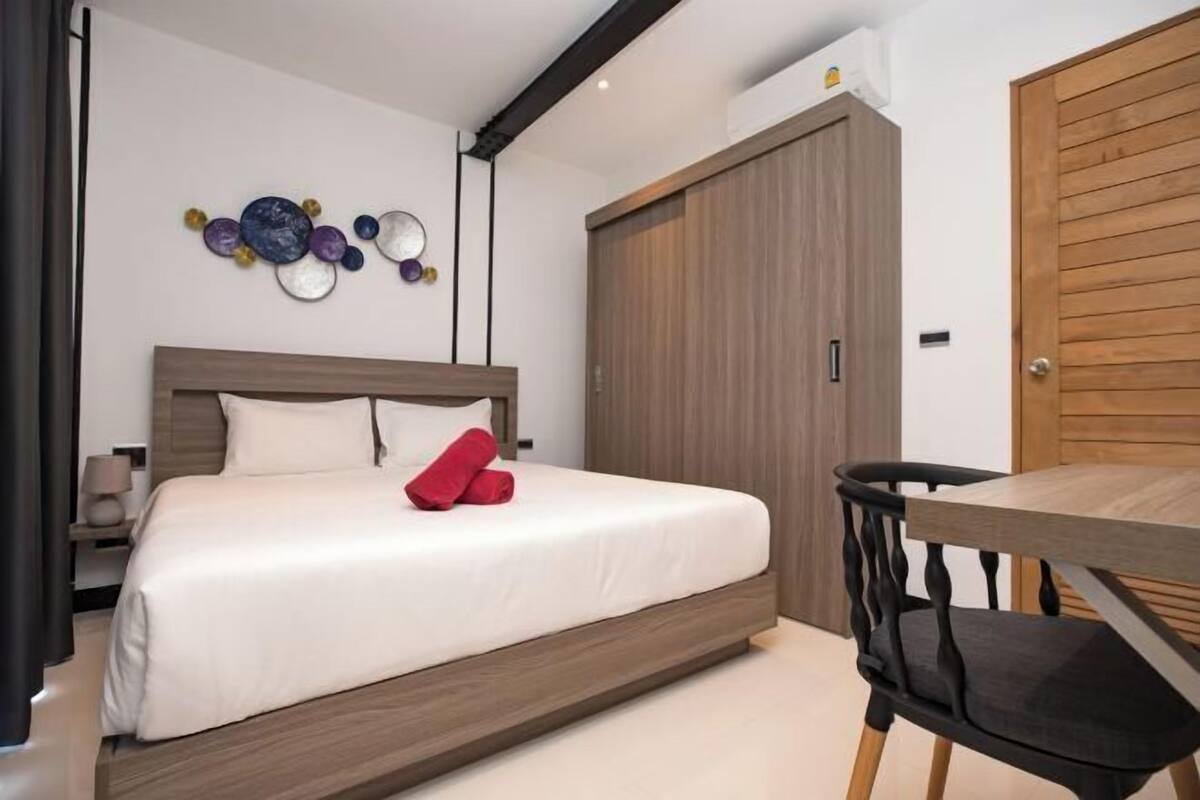 A bedroom with a bed, a wooden wardrobe, a dining table, and a chair.
