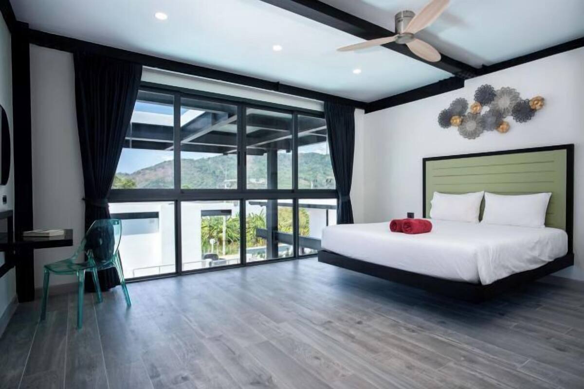 A modern bedroom with a large bed, a ceiling fan, and a view through floor-to-ceiling windows.