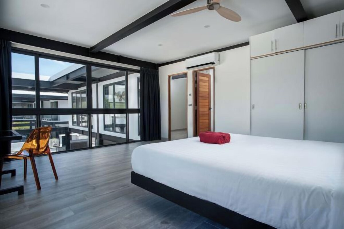 A modern bedroom with a large bed, a ceiling fan, and a view through a glass door to an outdoor area.