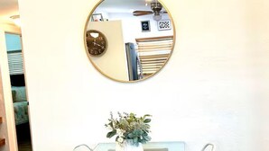 Uma área de refeições com mesa de vidro e cadeiras brancas, um espelho redondo na parede e uma pequena planta sobre a mesa.
