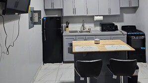 Eine kompakte Küche mit grauen Schränken, einem schwarzen Kühlschrank, einer Mikrowelle, einem Herd und einem Deckenventilator. Es steht ein kleiner Esstisch mit zwei Stühlen da, und an der Wand ist ein Fernseher angebracht.