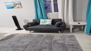 Ein modernes Wohnzimmer mit einem grauen Sofa, einem flauschigen grauen Teppich, einem kleinen weißen Beistelltisch, einer Fernbedienung und einem Gemälde mit einer Strandansicht.