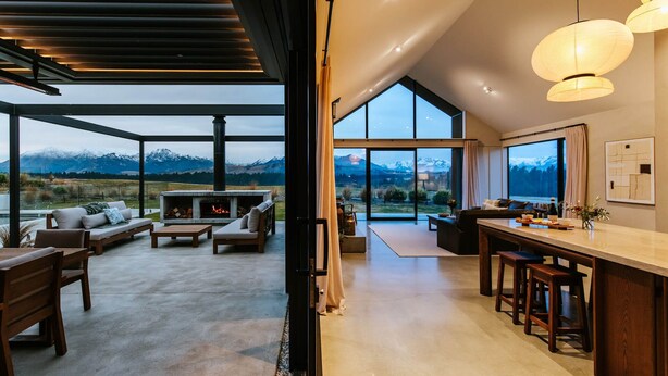 A modern interior with a large glass window overlooking mountains, a fireplace, and a dining area with bar stools. - Riverslea - Pool - Sleeps 10 (Wānaka)