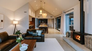 A modern living room with a fireplace, leather sofa, dining area, and kitchen in the background. - Riverslea - Pool - Sleeps 10 (Wānaka)