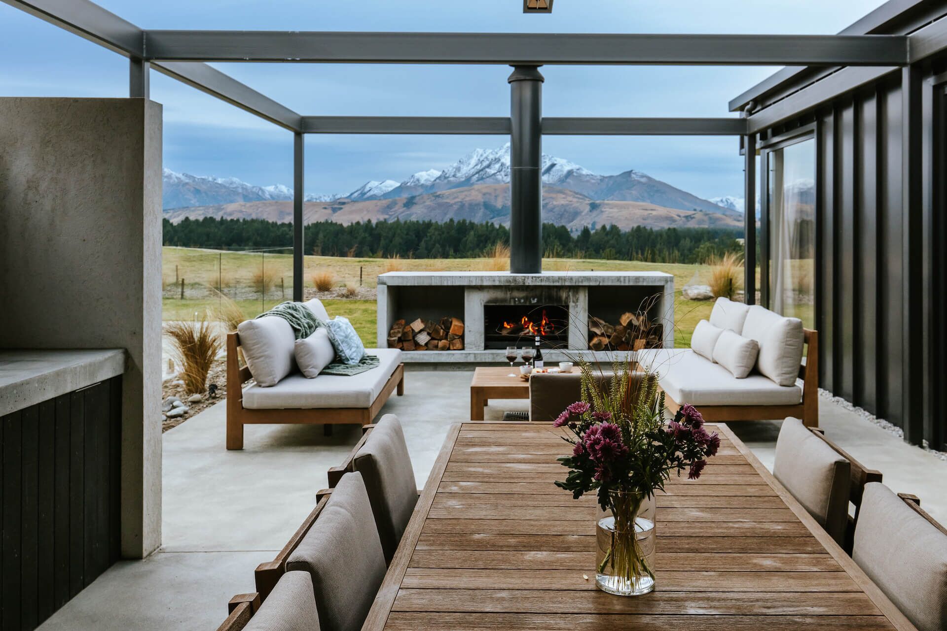 A covered outdoor seating area with a fireplace, sofas, and a wooden table.