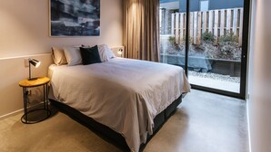 A modern bedroom with a large bed, a side table, a floor lamp, and a view of a garden through a sliding glass door. - Riverslea - Pool - Sleeps 10 (Wānaka)
