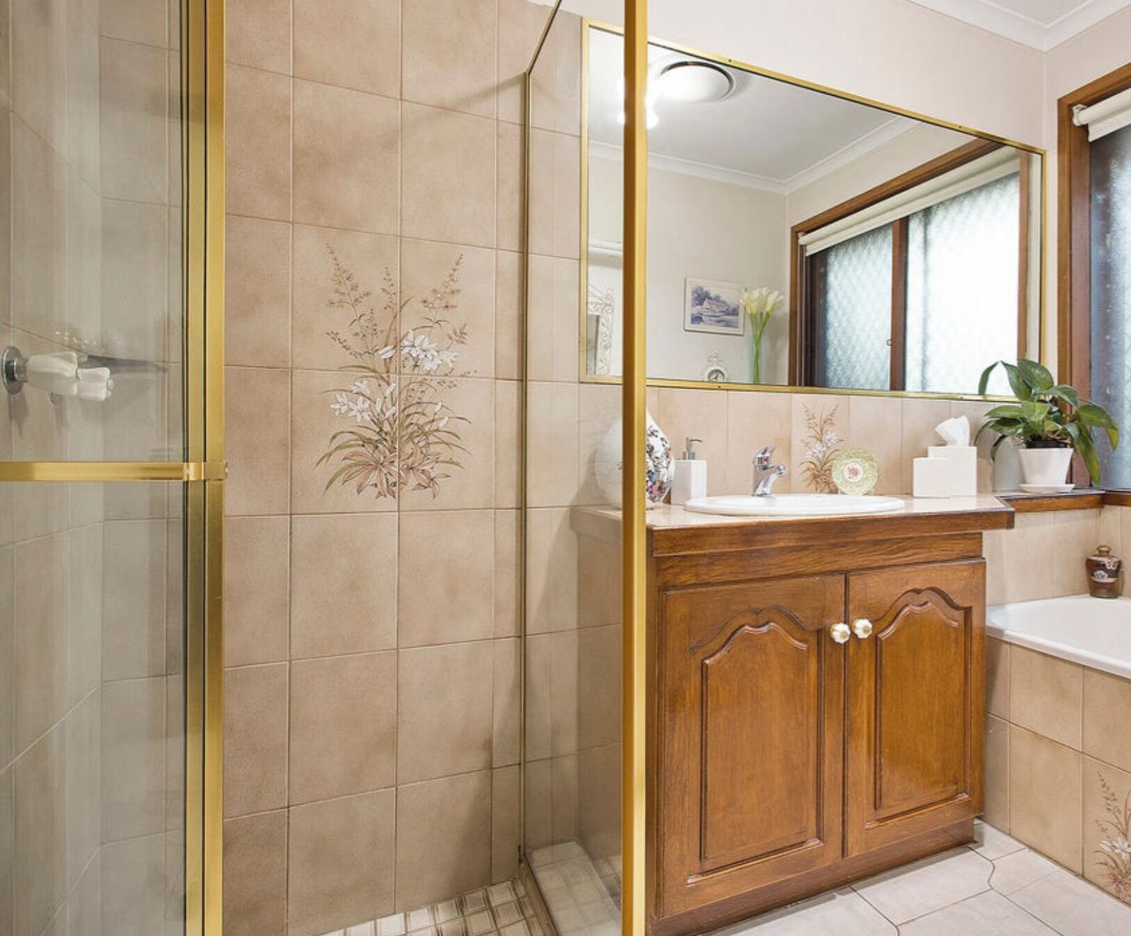 Une salle de bain avec une douche à parois de verre, des murs recouverts de carreaux beiges, un meuble-lavabo en bois avec un miroir et une plante en pot.