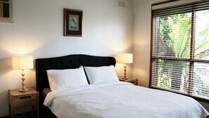 Una cama bien tendida con sábanas blancas, dos mesitas de noche con lámparas, un cuadro en la pared y una ventana con persianas.