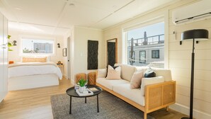 Moderni hotellihuone, jossa on sänky, sohva, sohvapöytä ja suuri ikkuna, josta avautuu näkymä kaupunkiin.