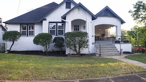 A white two-story house with a dark roof, a front porch, and a small front yard with trimmed bushes.
