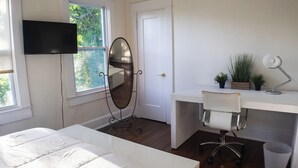 A bedroom with a bed, a desk with a chair, a mirror, a TV, and a window with a view of trees.