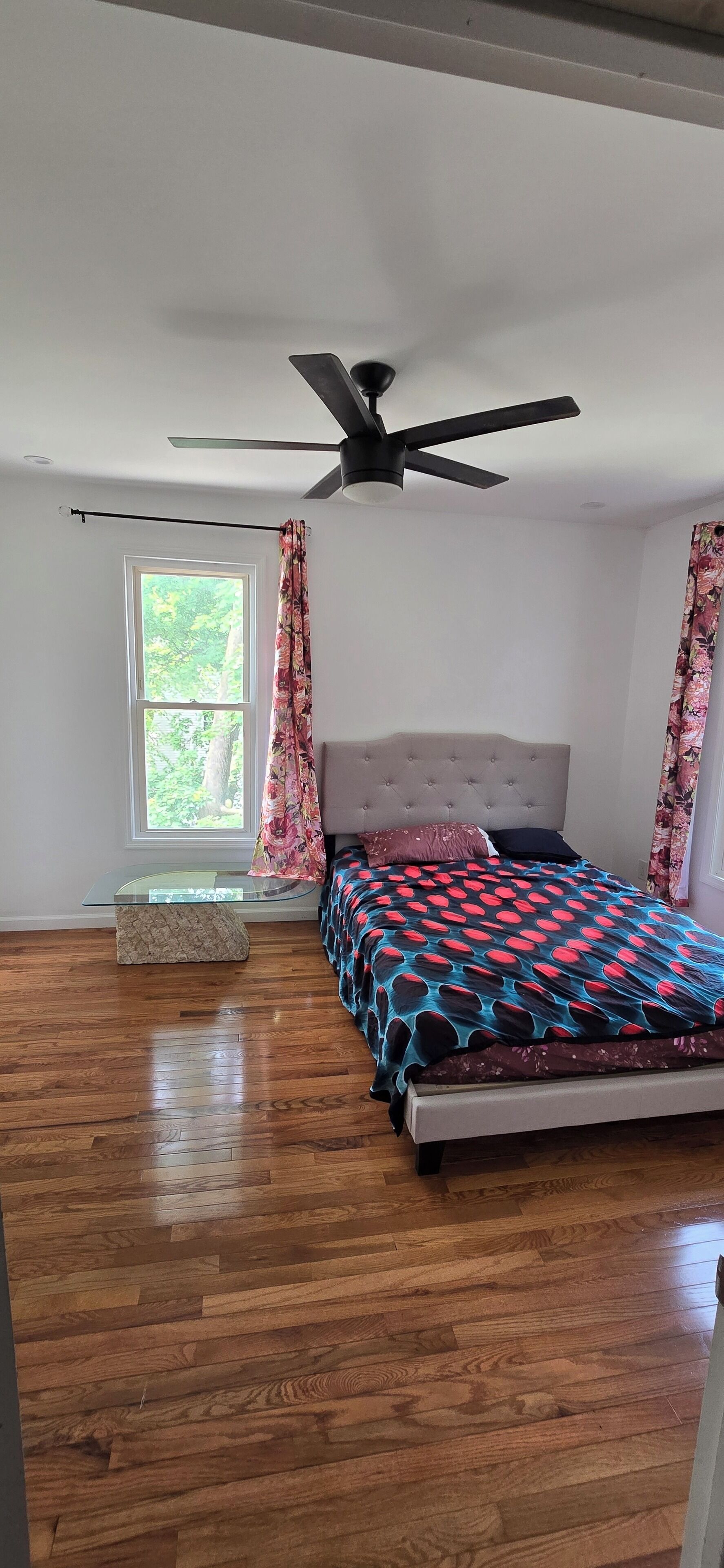 Une chambre à coucher avec un lit, un ventilateur de plafond, une fenêtre avec des rideaux et un plancher en bois.