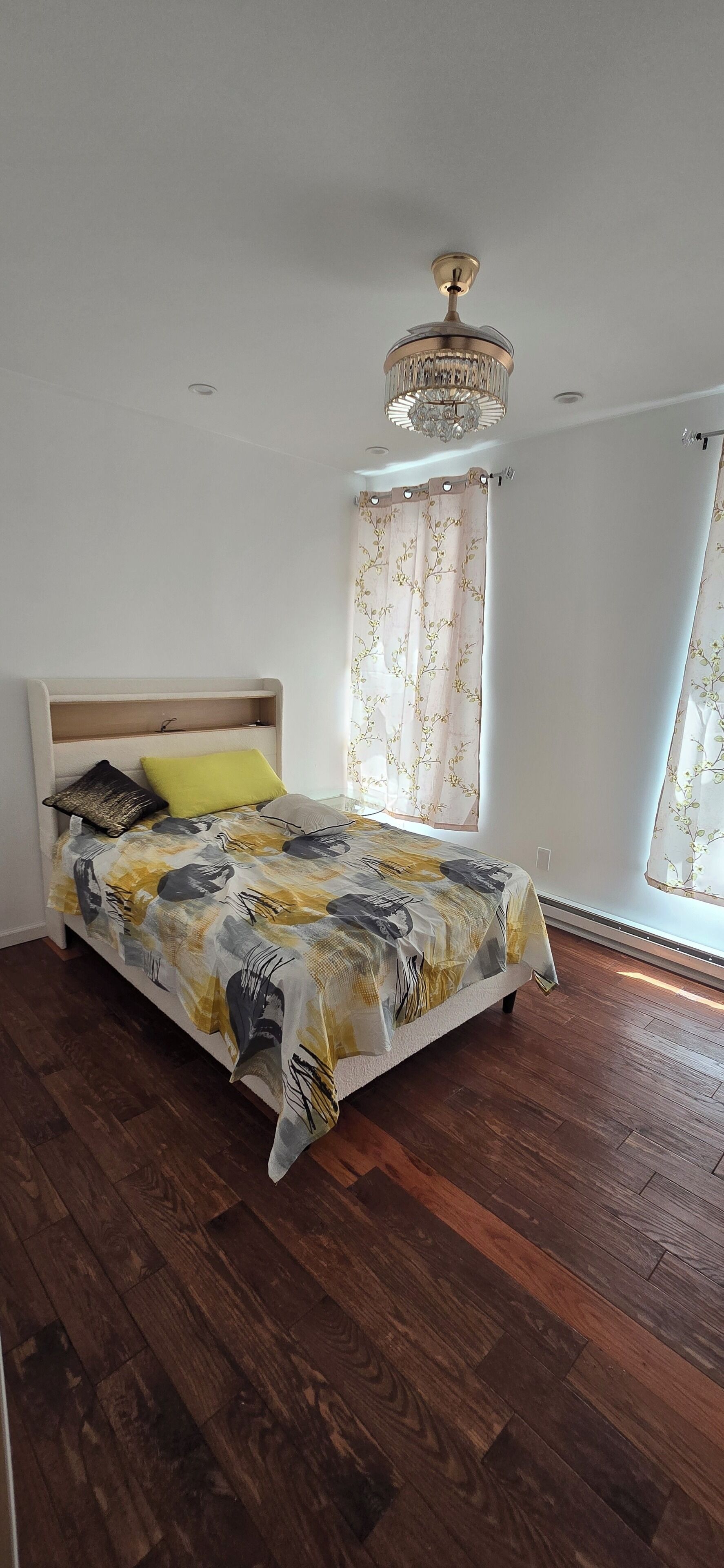 A bedroom with a bed, wooden flooring, a chandelier, and a window with curtains.
