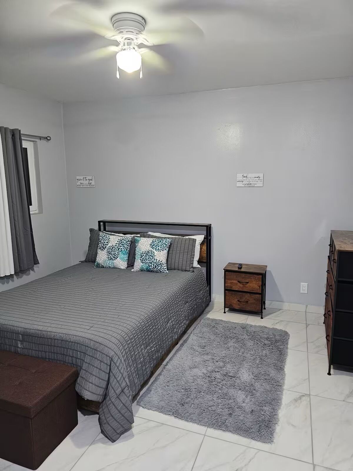 A bedroom with a bed, a nightstand, a dresser, a ceiling fan, and two wall decorations.