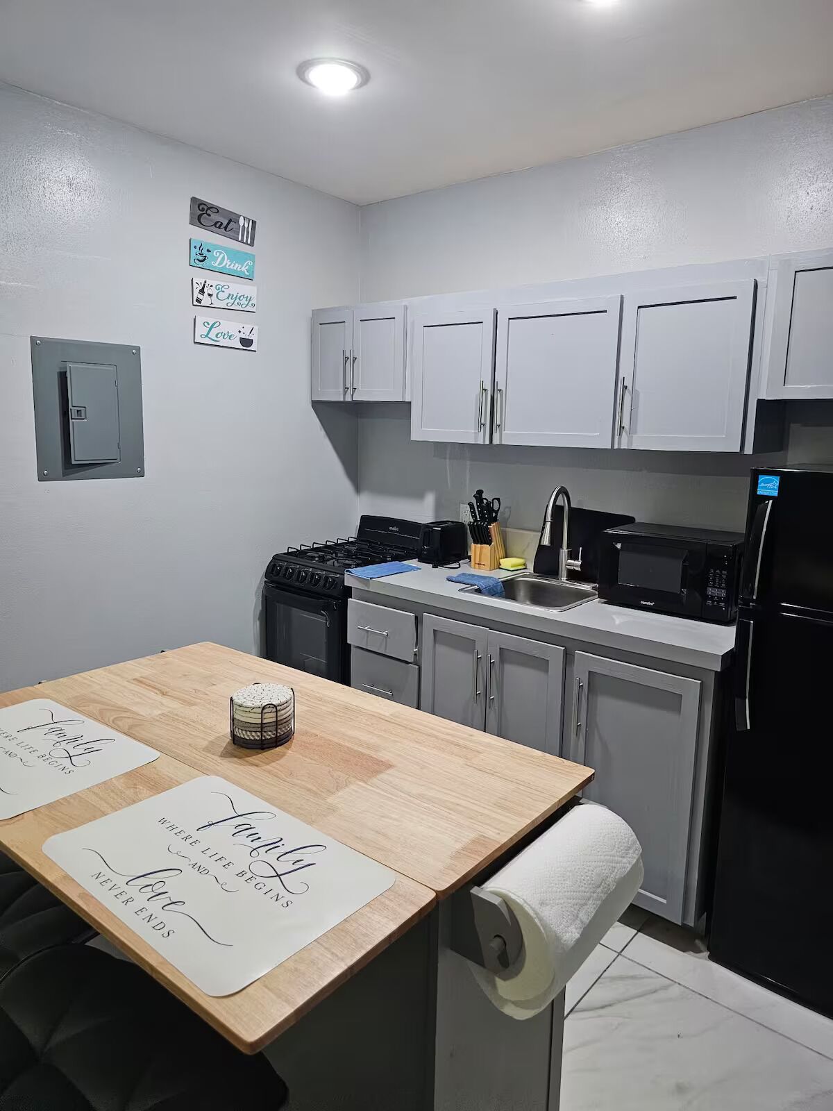A kitchen with white cabinets, a stove, a sink, and a refrigerator. There are signs on the wall and a table with a roll of paper towels.