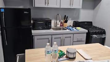 A modern kitchen with grey cabinets, a black refrigerator, a microwave, a sink, and a stove. There are two water bottles on the counter, a sponge, and a welcome mat with a family-themed message.