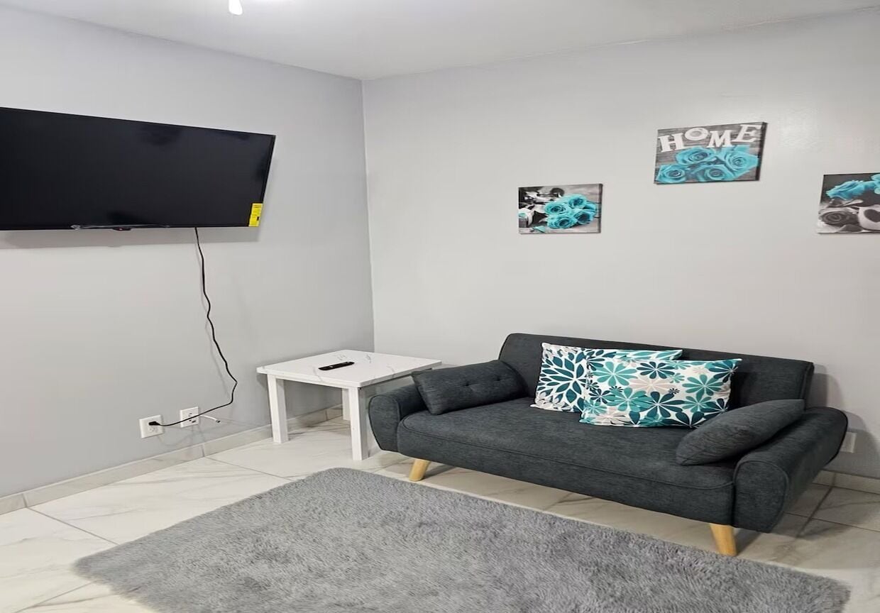 A modern living room with a grey sofa, a flat-screen TV mounted on the wall, a small white side table, and a grey rug.