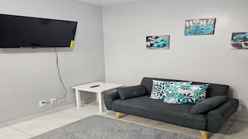 A modern living room with a grey sofa, a flat-screen TV mounted on the wall, a small white side table, and a grey rug.