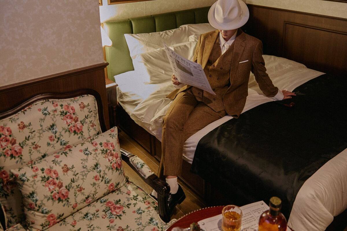 Una persona sentada en una cama leyendo un libro en una habitación con un sillón de estampado floral y cabecera de madera.