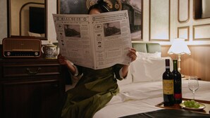 Eine Person liest eine Zeitung in einem Hotelzimmer, mit Radio, Weinflasche und Glas auf dem Nachttisch.